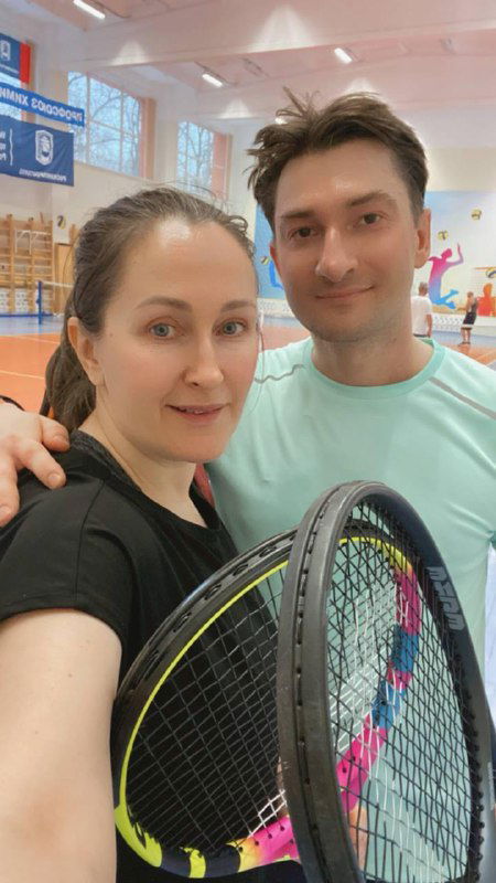 Селфи двух взрослых после тренировки: люди держат теннисные ракетки в спортивном зале, на заднем плане виден корт и освещение.