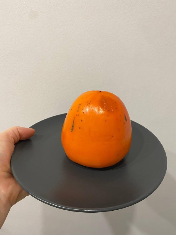 Хурма, аккуратно поданная на тёмной тарелке, крупный план фрукта на нейтральном фоне, натюрморт в домашней обстановке.