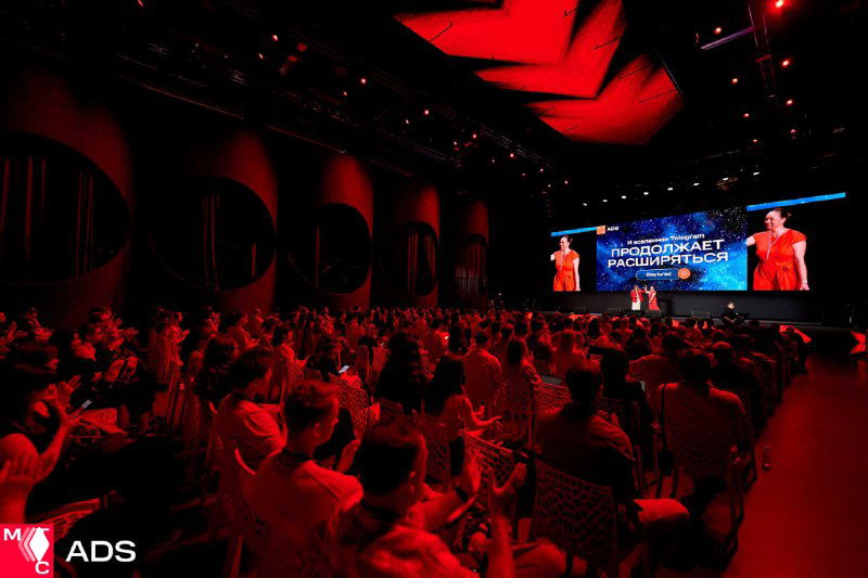 Зал с аудиторией во время сессии конференции: люди следят за выступлением на большом экране при сценическом освещении.