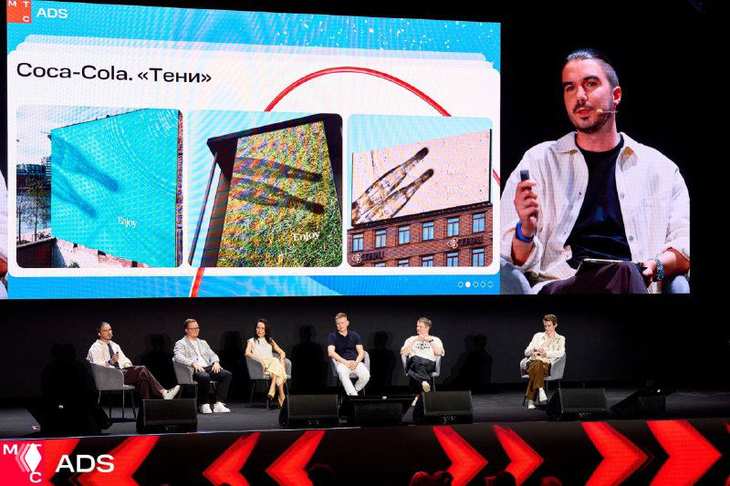 Панель спикеров на сцене: несколько экспертов сидят в ряду, крупный экран за ними демонстрирует слайды, публика внизу.