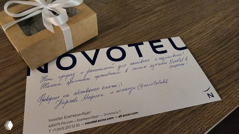 Приветственная открытка с логотипом Novotel и небольшая подарочная коробочка с конфетами на деревянной поверхности в номере отеля, видна подпись и оформление.