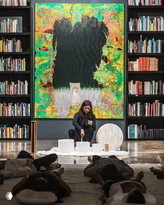 Внутренний зал музея: человек сидит перед большим ярким полотном, участники лежат на ковриках — подготовка к групповому фитнес-занятию в галерее от Equinox.