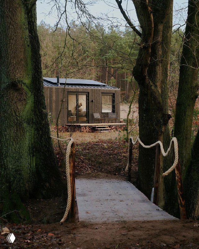 Маленький модульный домик-глэмпинг за деревянным настилом и канатным перилами, окружённый лесной подлеской и тропой к входу.