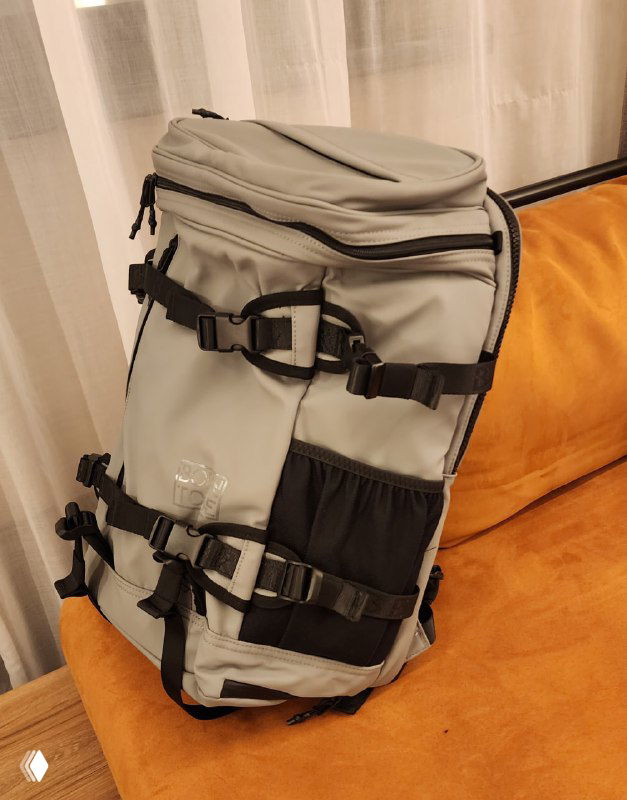 Серый городской туристический рюкзак с множеством ремней, застёжек и карманов, лежащий на оранжевом диване рядом со шторами — фото из поста.