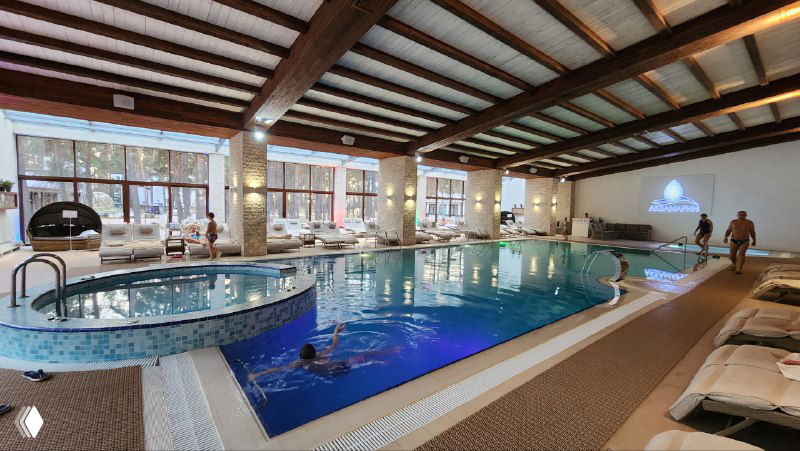 Интерьер спа-центра с крытым бассейном и джакузи, шезлонгами у воды, панорамными окнами и отдыхающими гостями в курортной атмосфере.