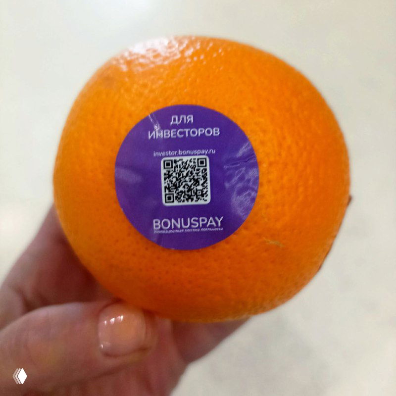 Рука держит мандарин с фиолетовой круглой наклейкой «для инвесторов», на ней QR‑код и надпись BONUSPAY — пример промо с QR на выставке.