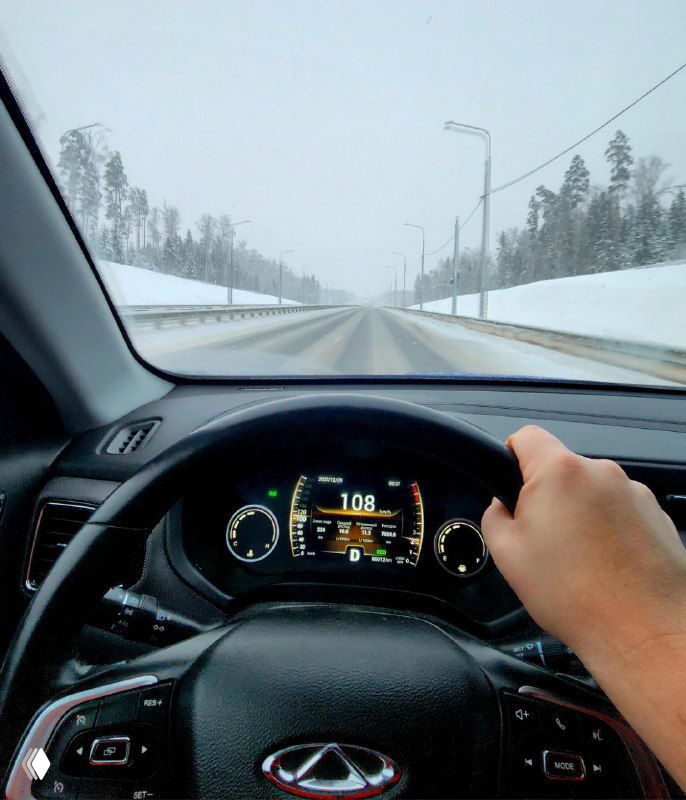 Вид из салона автомобиля на заснеженную дорогу во время метели: рулевое колесо, приборная панель и правая рука водителя, ощущение зимнего путешествия.