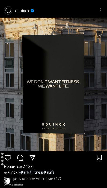 Черный наружный рекламный щит на фасаде здания с английским слоганом «WE DON'T WANT FITNESS. WE WANT LIFE», визуал указывает на кампанию сети фитнес-клубов в городском контексте.
