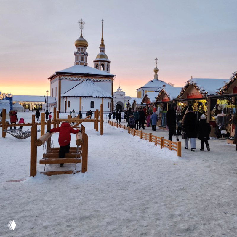 Ряд сувенирных лавок у стены исторического ансамбля, люди заходят и греются, вокруг деревянные ограждения и снежная дорожка — рождественский базар.