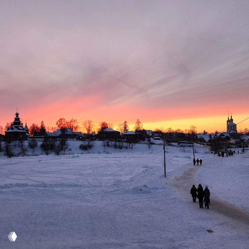 Широкая панорама заснеженного ландшафта Суздаля на фоне огненного заката; внизу несколько фигур людей, идущих по дороге — атмосферный пейзаж.