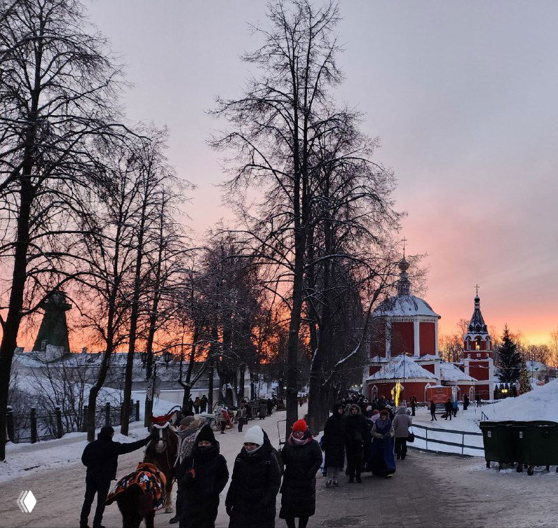 Снова людная пешеходная аллея Суздаля на закате, видны купола и праздничные украшения, люди толпятся у туристических точек — живой городской кадр.