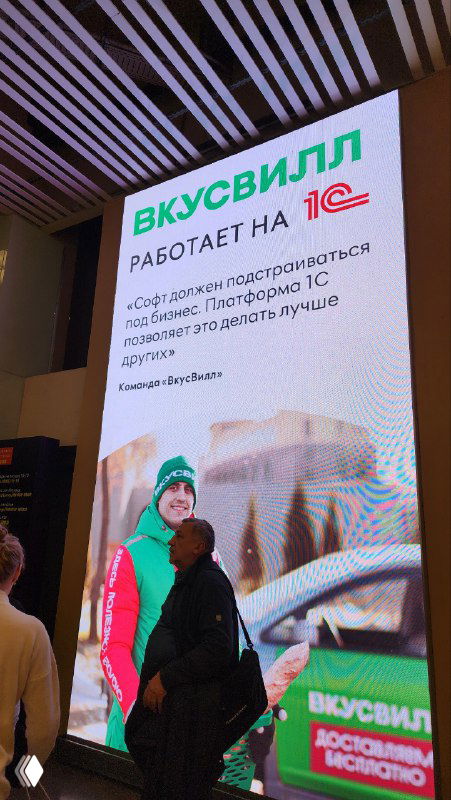 Фото крупного наружного баннера в аэропорту: логотип «Вкусвилл», надпись о работе на 1С и прохожие на фоне, пример кобрендинга.