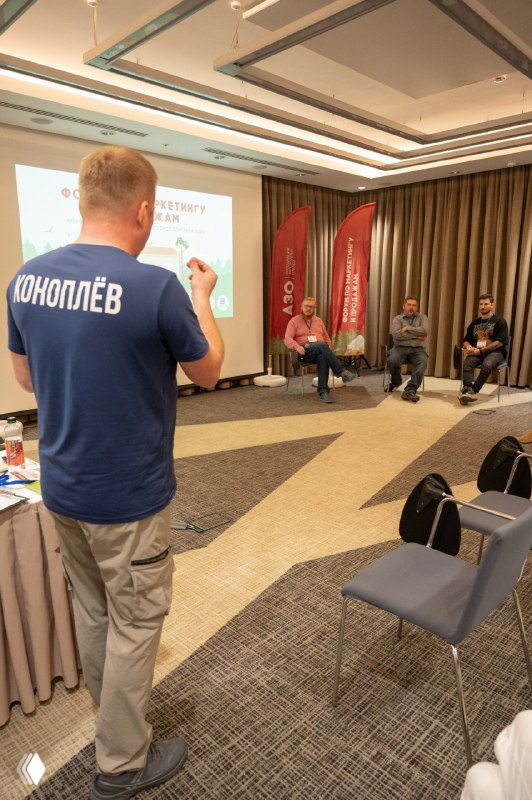 Человек в футболке с надписью «КОНOПЛЁВ» на фоне сцены и зала конференции: доклад на форуме загородных отельеров, проектор и участники в зрительном зале.