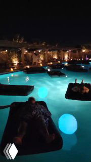 Ночное изображение: люди на матрасах плавают в подсвеченном бассейне курорта, создавая атмосферу уединённого велнес-опыта.
