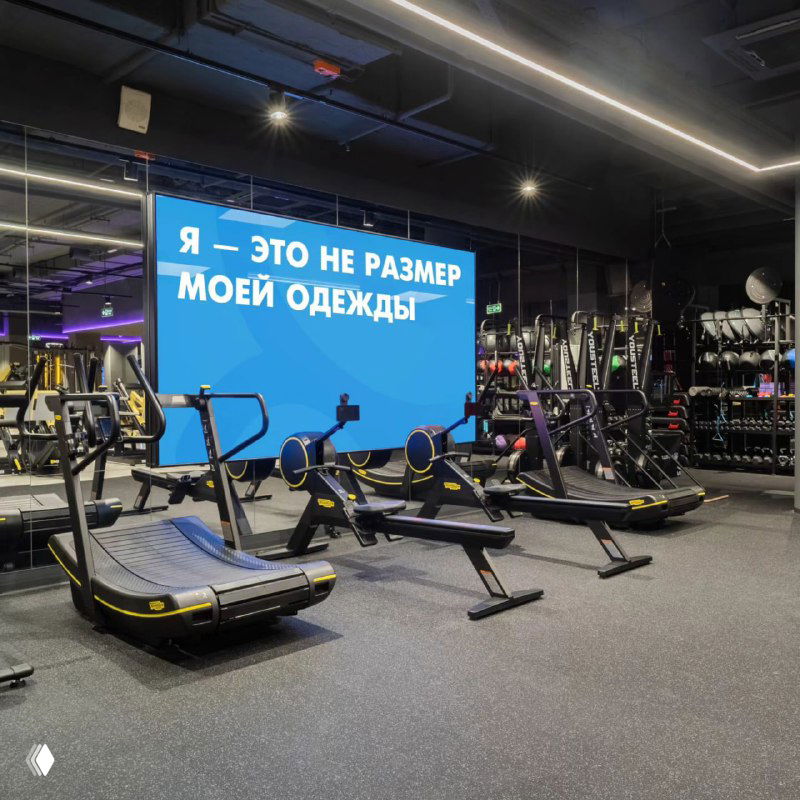 Интерьер современного фитнес‑клуба DDX с кардиооборудованием и большим экраном, на котором виден слоган о самооценке и теле клиента.