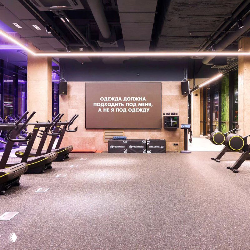Пустой зал с тренажерами и большим экраном, на котором размещён месседж о том, что одежда должна подходить человеку, а не наоборот.