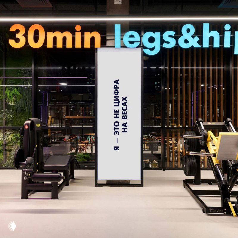 Тренажёрный зал с силовыми тренажерами и неоновым заголовком «30min legs&hip», а также вертикальным экраном с коротким тезисом о весах.