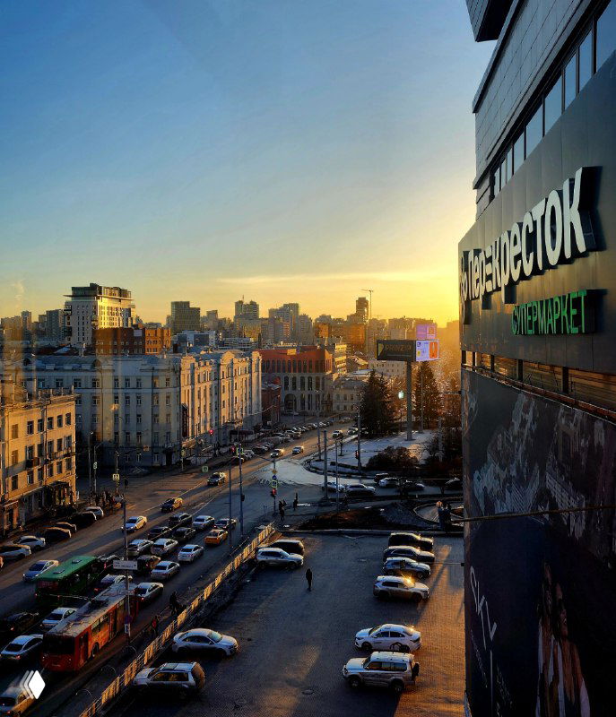 Панорамный вид Екатеринбурга с высоты: улица с припаркованными машинами и трамваем, вечернее небо и фасад с вывеской.