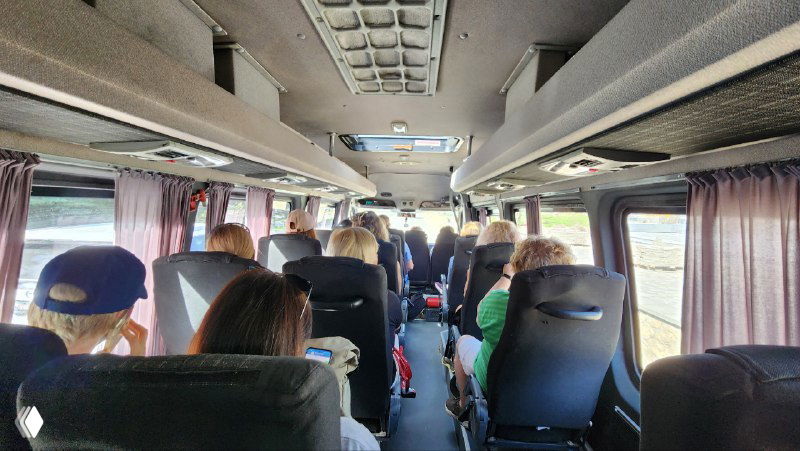 Интерьер автобуса с сидящими участниками кэмпа, едущими на активность; видны пассажиры, сиденья, проход и занятые места в салоне.