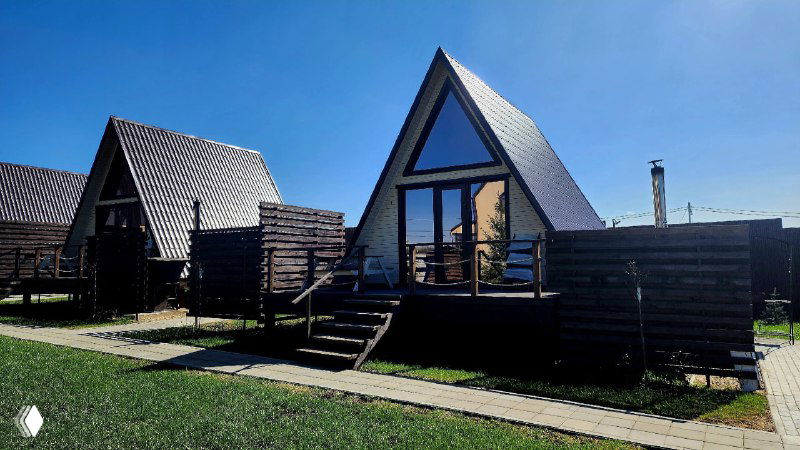 Деревянные дачные домики Pavlov Village на зелёной лужайке при ясной погоде, вид фасадов, террас и аккуратной центральной аллеи рядом с газонами.