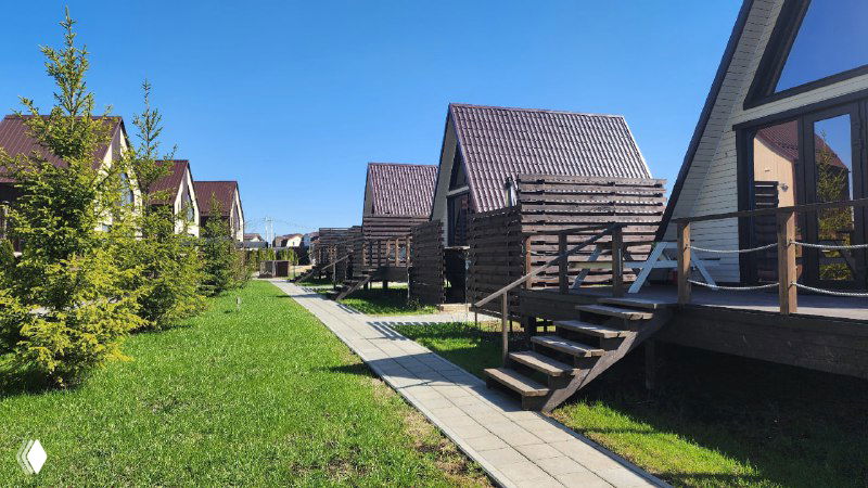 Аллея между коттеджами Pavlov Village: аккуратные тротуары, террасы домиков и молодые деревья на фоне чистого неба, вид вдоль застроенной зоны.