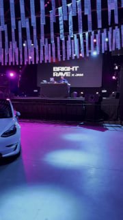Сцена Rave Night с подсветкой и инсталляцией BRIGHT RAVE, на переднем плане припаркована белая Tesla и видны фирменные брендированные гоночные машины в концертном холле.