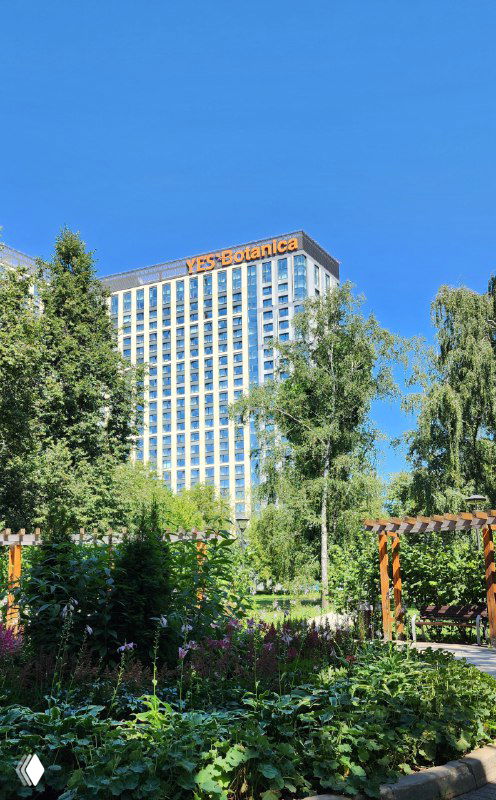 Фасад многоэтажного отеля в Москве за зеленью ботанического сада: высокое здание с вывеской и летним садом на переднем плане.