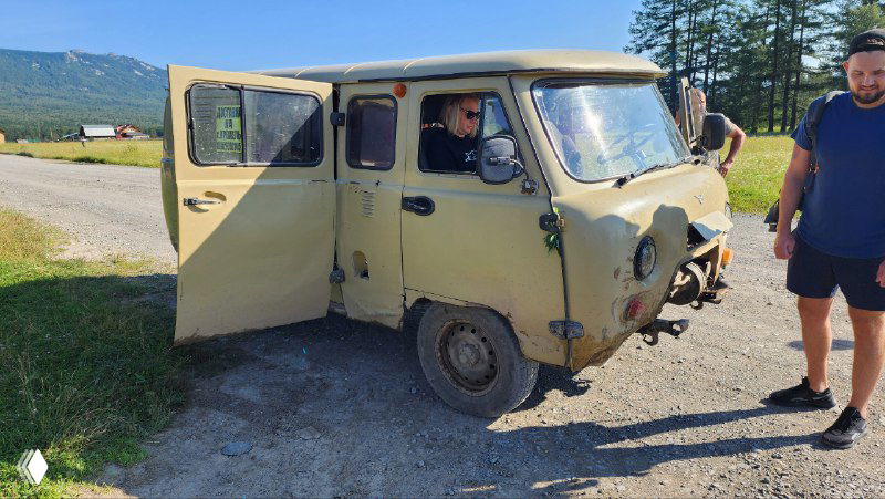 Жёлтый микроавтобус «буханка» с открытой дверью на просёлочной дороге у подножия горы; рядом туристы и водитель.