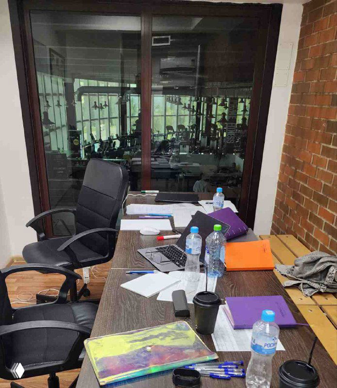 Офисный стол в фитнес-центре: ноутбук, папки, бутылки воды и кружка на столе; за стеклом виден тренажёрный зал с оборудованием и кардиозоной.