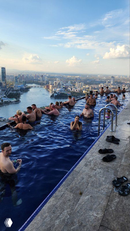 Участники в видовом бассейне на крыше высотного здания: купающиеся и фотографирующиеся на фоне городской панорамы и реки.