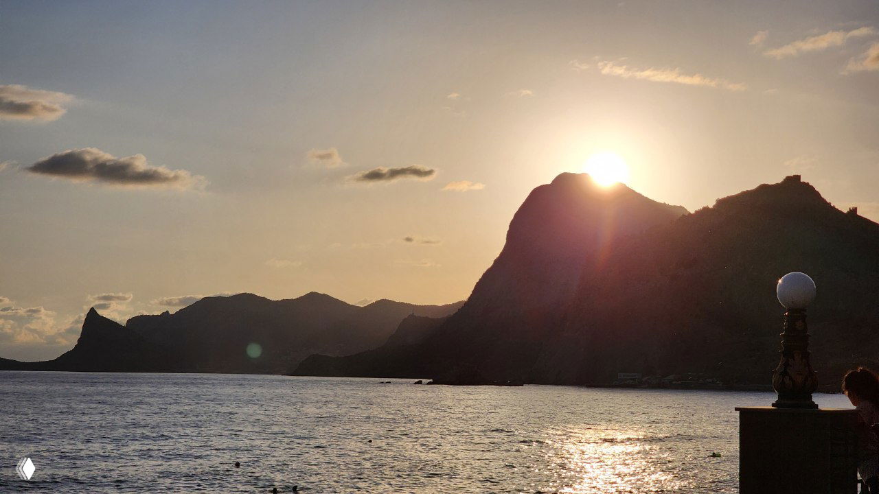 Закат над морем в Судаке: силуэты гор на горизонте, блеск заходящего солнца на воде и пустая прибрежная набережная в низкий сезон.