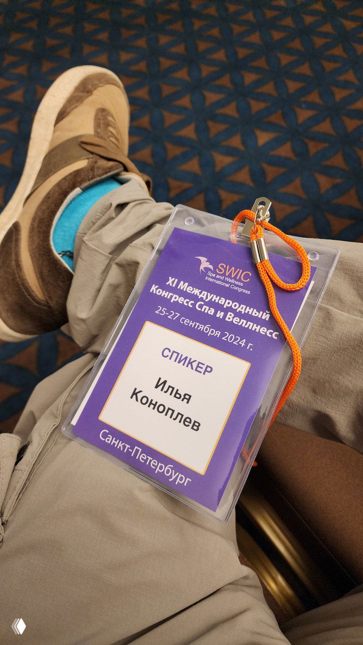 Бейдж спикера «СПИКЕР Илья Коноплёв» на коленях, оранжевый шнурок, на фоне кресел и ковра в зале конгресса.