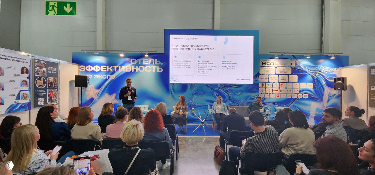 Передняя часть зала и сцена на выставке: докладчик у экрана на фоне брендированного баннера «Отель. Эффективность», аудитория в креслах.