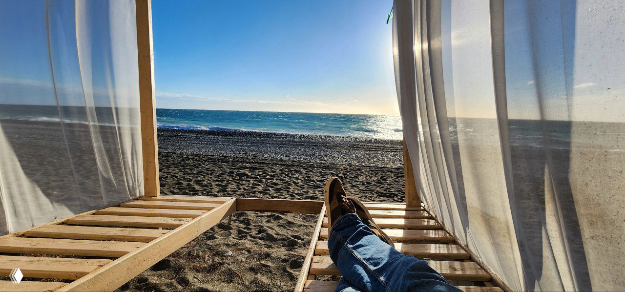 Вид на морской берег из пляжной беседки: ноги человека в джинсах на лежаке, песок и спокойное море на рассвете — иллюстрация сценария уединённого отдыха.