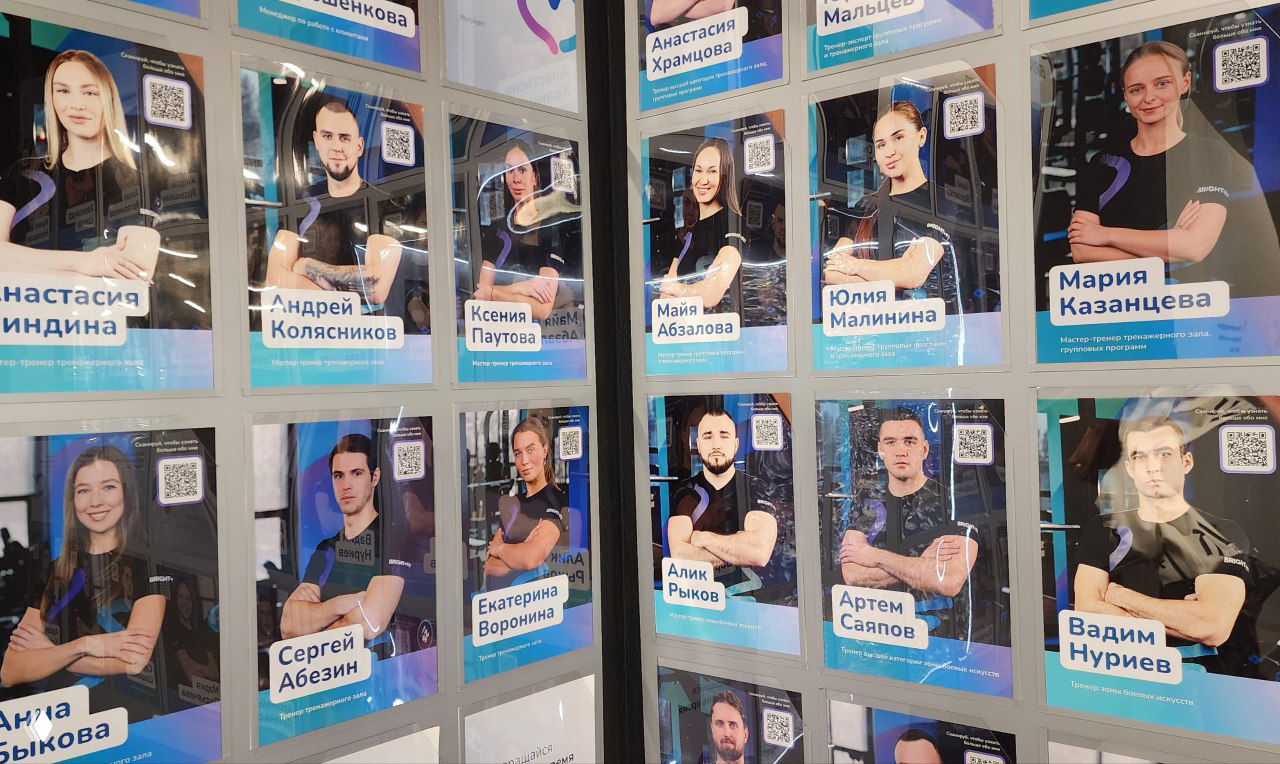Фотографии и постеры персональных тренеров на стенде фитнес‑сети с портретами и QR‑кодами для перехода на их страницы в приложении.
