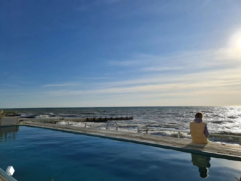 Открытый бассейн у моря в Сочи: человек сидит на бортике, вид на море и широкое ясное небо в декабре, спокойная атмосфера курорта.