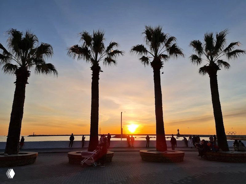 Набережная Сочи на закате: стройные пальмы, лежаки и силуэты людей на фоне оранжевого горизонта у моря, тёплые краски вечера.