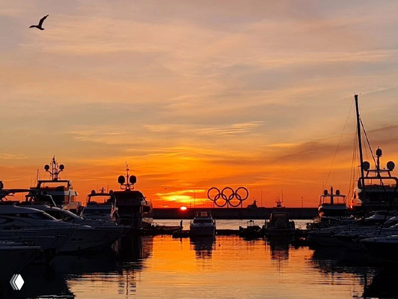 Яхтенный причал в Сочи на закате: лодки отражаются в воде, оранжевое небо и силуэты причала с заметными Олимпийскими кольцами на фоне.