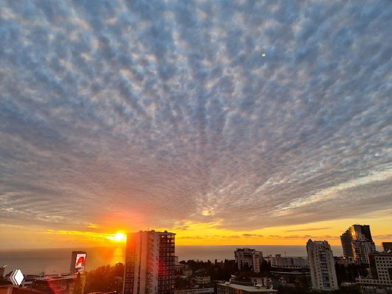 Широкий вид на закат в Сочи с высоты: плотные облака, огни города и море вдалеке, тёплые оттенки неба и городской силуэт.