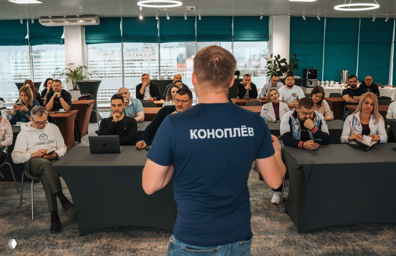 Спикер в футболке с надписью «КОНОПЛЁВ» выступает перед аудиторией на конференции в Сочи; участники слушают, работают на ноутбуках и делают заметки.