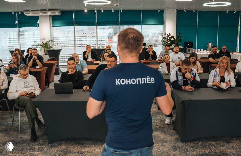 Ведущий спиной в синей футболке перед аудиторией; участники на конференции сидят за столами с ноутбуками и блокнотами в делово-wellness формате события.