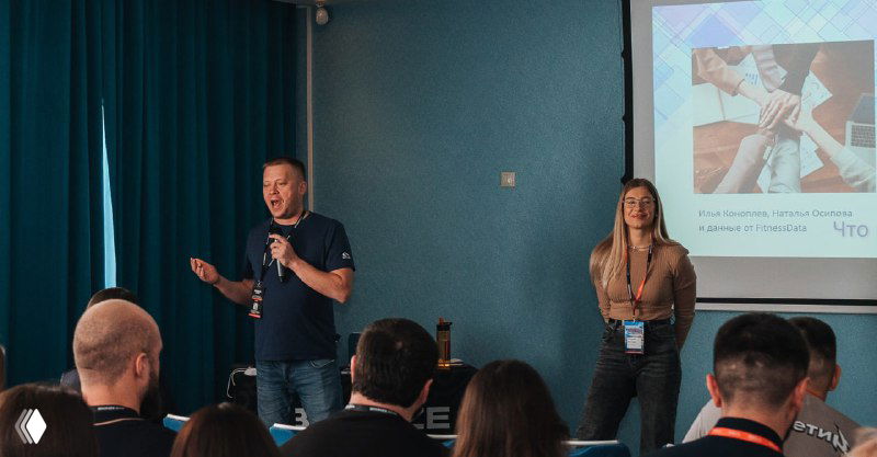 Спикер и ведущая на сцене в конференц-зале перед аудиторией; видно проектор, экраны и деловую атмосферу мероприятия для организаторов.