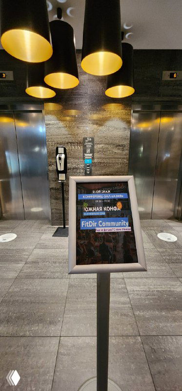 Пустая фойе зона с указателем и стойкой регистрации у входа в конференц-зал, кадр отражает логистику и навигацию на мероприятии.