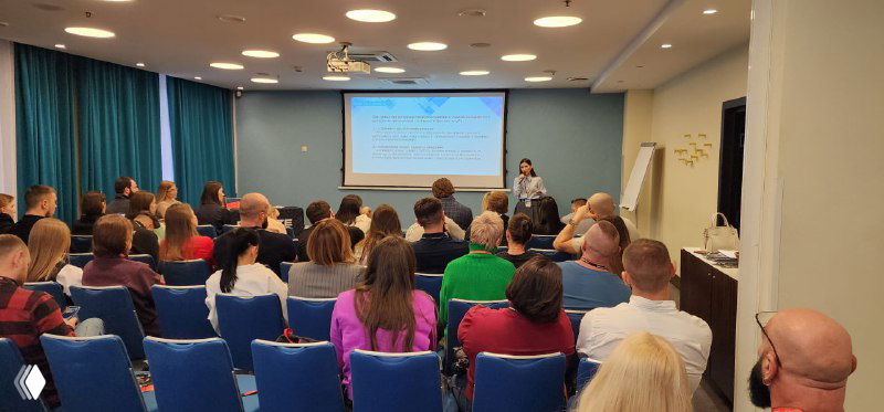 Спикер выступает перед полной комнатой участников; видно экран с презентацией и участников, участвующих в дискуссии.