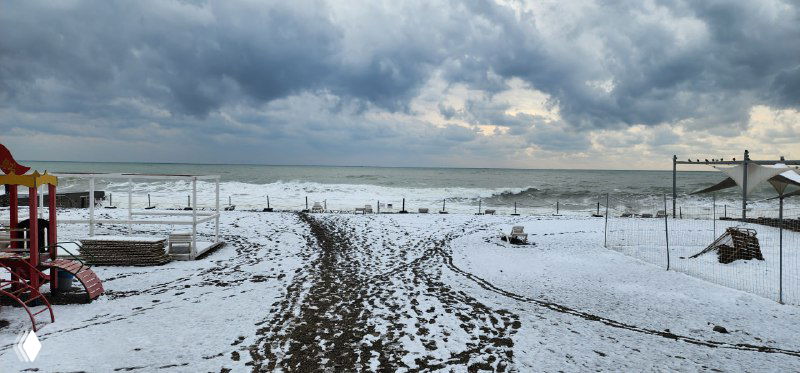 Заснеженный пляж в Сочи: следы на песке, пустые шезлонги и зонтики на фоне серого пасмурного неба и моря, вид из кэмпа.