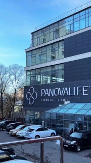 Фасад коммерческого здания с крупной вывеской фитнес‑клуба PanovaLife, перед входом припаркованы автомобили, городской дневной пейзаж и стеклянный вестибюль.
