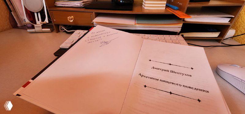 Фотография открытой книги с рукописным пожеланием автора на внутренней обложке и рабочим столом на фоне.