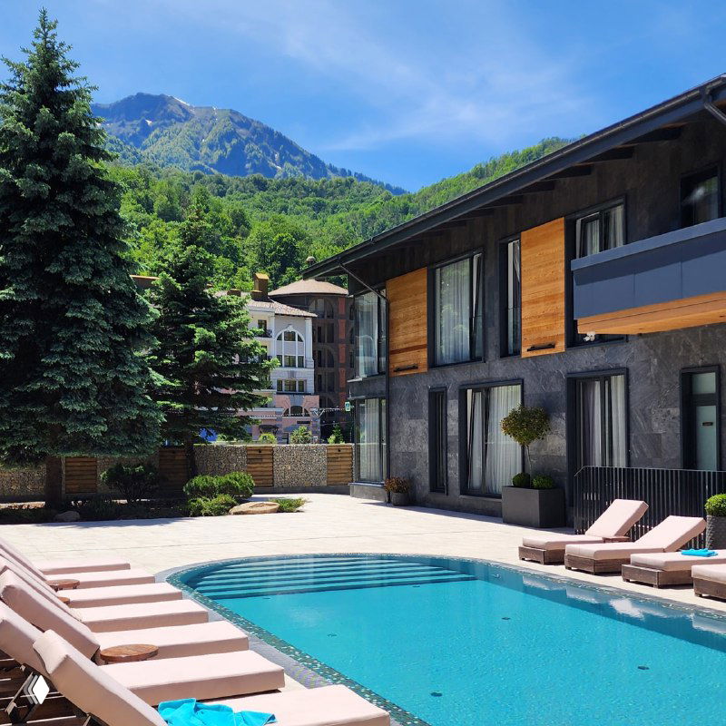 Вид на бассейн отеля Blanc Hill на фоне гор: шезлонги у воды, хвойные деревья, корпуса курорта и ясное голубое небо, создающие атмосферу, от которой не хотелось уезжать.