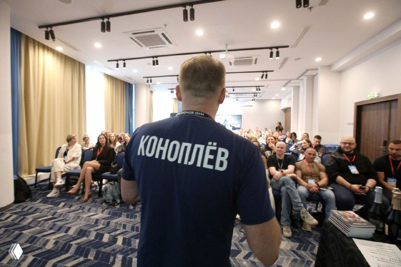 Ведущий конференции в футболке с надписью «КОНОПЛЁВ» на фоне аудитории: спич, презентация и живой зал участников.