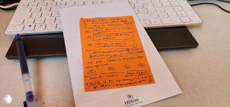 Фото рабочего стола: оранжевый стикер с рукописными заметками на бланке отеля (Hilton), рядом ручка и клавиатура — план проекта ивента.
